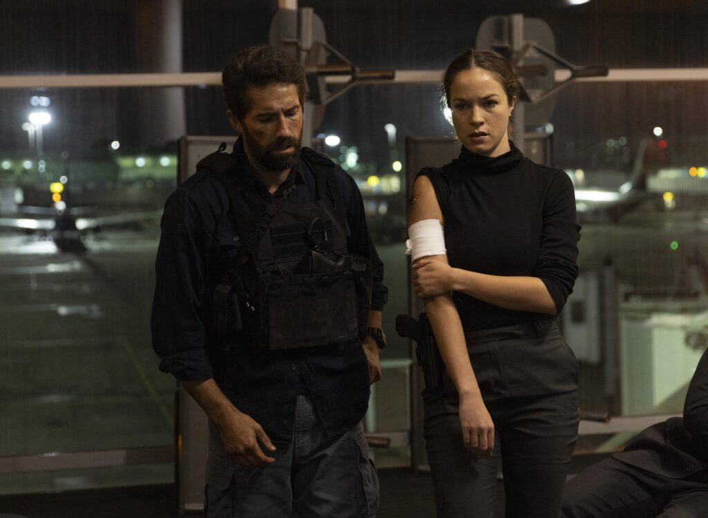 A young man and a woman in dark clothes and serious looks on their face, the woman holding her bandaged arm