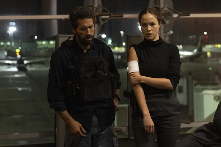 A young man and a woman in dark clothes and serious looks on their face, the woman holding her bandaged arm