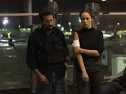A young man and a woman in dark clothes and serious looks on their face, the woman holding her bandaged arm