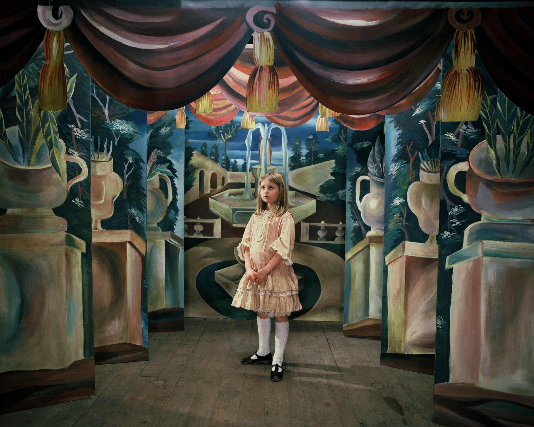 A little girl in a pink dress standing, looking lost, in a hallway with paintings on the walls