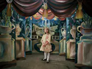 A little girl in a pink dress standing, looking lost, in a hallway with paintings on the walls