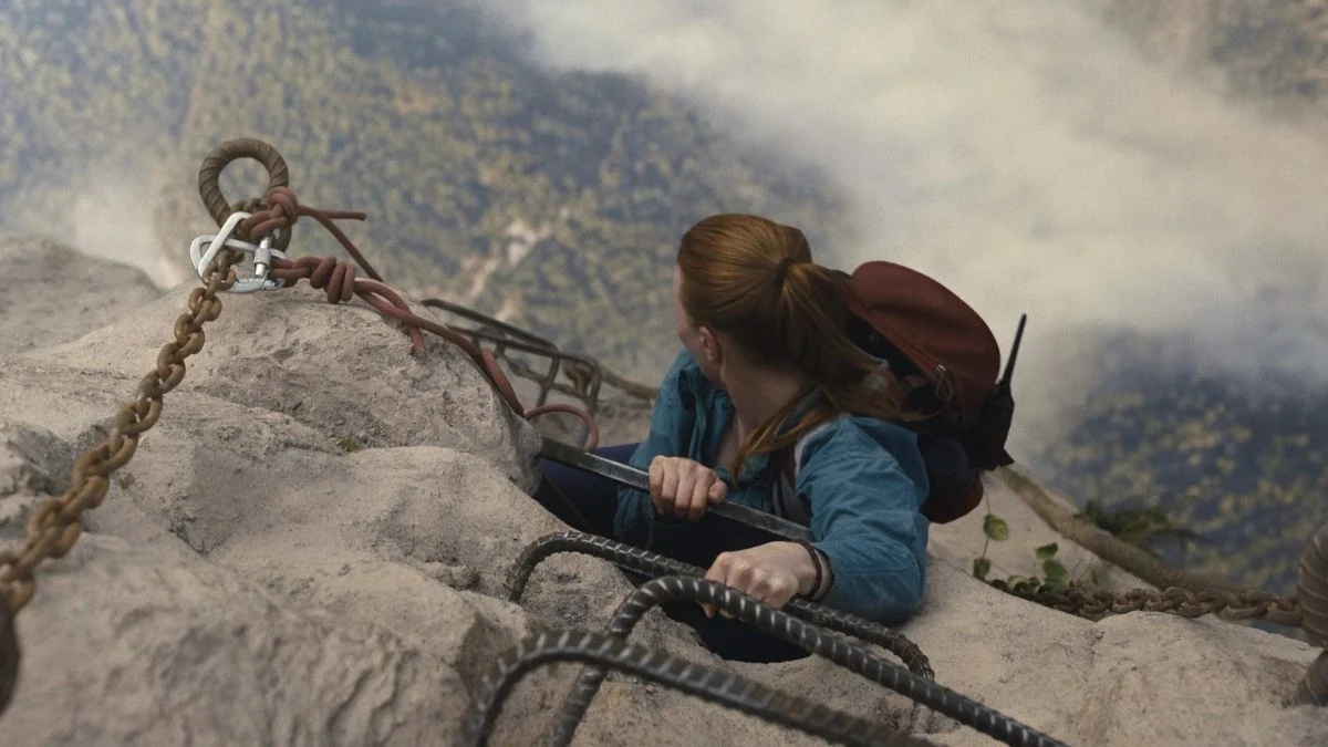 An aerial shot of a red-haired woman hanging on to the edge of a mountain, looking down at the ground far down