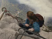 An aerial shot of a red-haired woman hanging on to the edge of a mountain, looking down at the ground far down