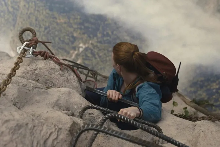 An aerial shot of a red-haired woman hanging on to the edge of a mountain, looking down at the ground far down