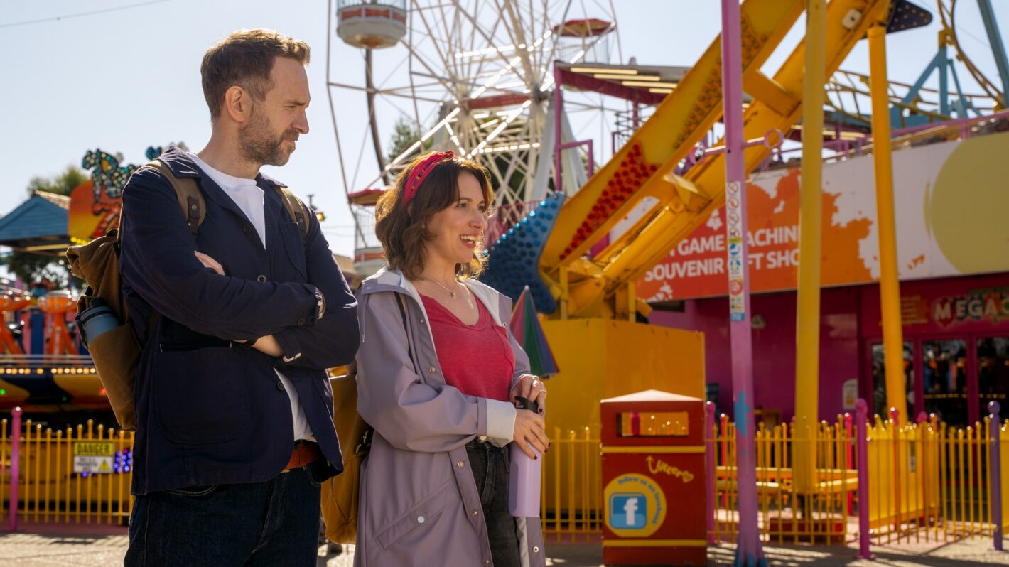 A white man and white woman wearing light jackets walking past a yellow carousel at a carnival