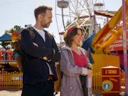 A white man and white woman wearing light jackets walking past a yellow carousel at a carnival