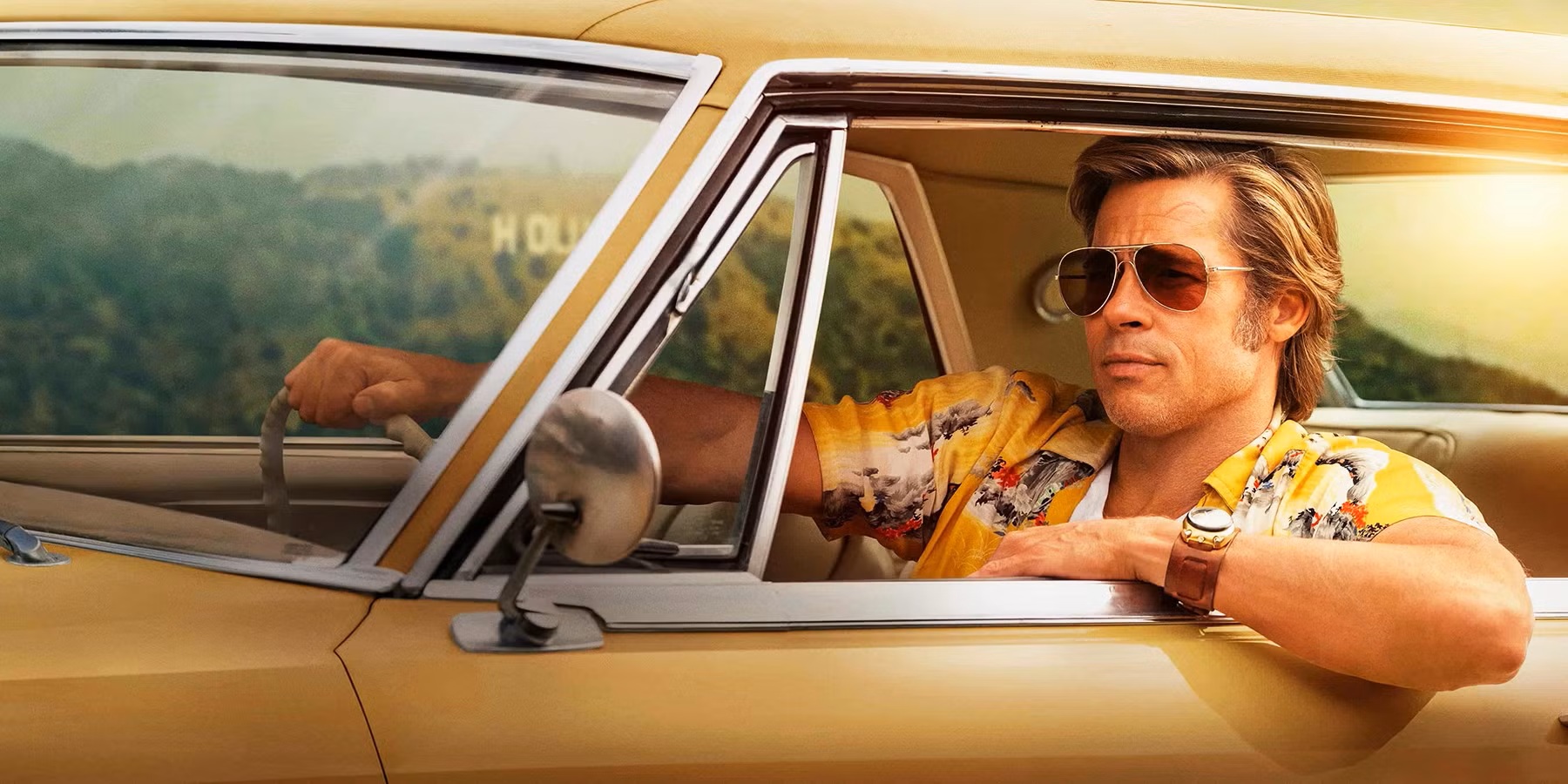 A man wearing sunglasses and a yellow shirt driving in Hollywood with his elbow out the window. The sun is behind him