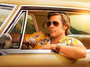 A man wearing sunglasses and a yellow shirt driving in Hollywood with his elbow out the window. The sun is behind him