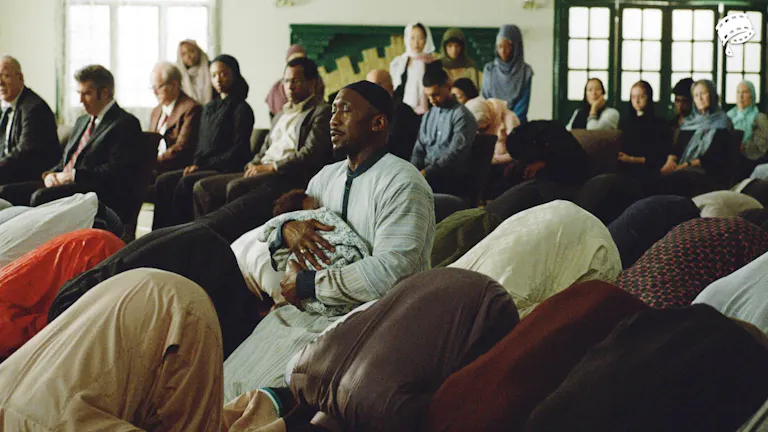 A man kneeling in a mosque, eyes closed in prayer while holding a baby in his arms. Around him are other men praying with their faces to the ground