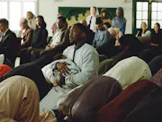 A man kneeling in a mosque, eyes closed in prayer while holding a baby in his arms. Around him are other men praying with their faces to the ground
