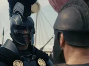 A close up of a man wearing blue ancient war armour, with a large ship in the background