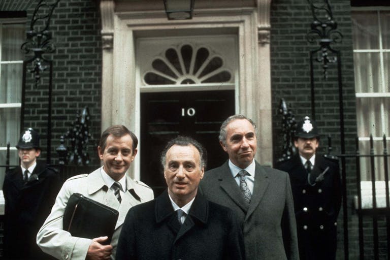 Three white middle aged men in suits standing in front of the door to 10 Downing Street flanked by two police officers