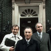 Three white middle aged men in suits standing in front of the door to 10 Downing Street flanked by two police officers