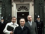 Three white middle aged men in suits standing in front of the door to 10 Downing Street flanked by two police officers