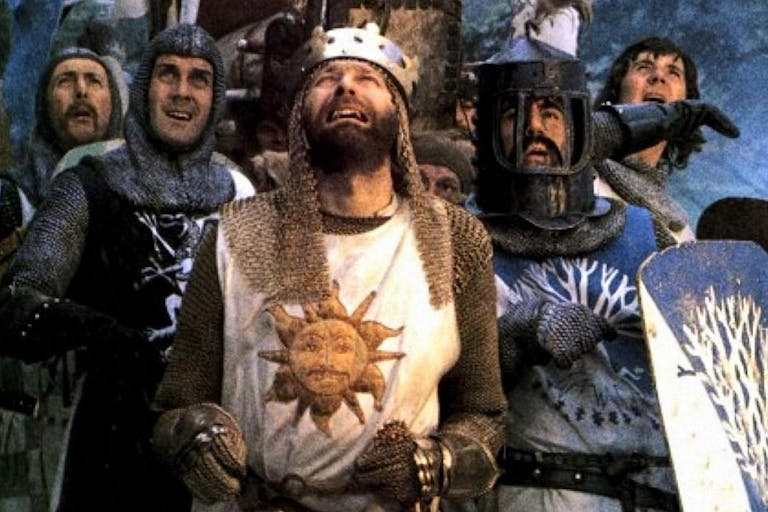 Five white men in chainmail and holding shields, one wears a crown, they all look up in comical distress