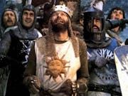 Five white men in chainmail and holding shields, one wears a crown, they all look up in comical distress