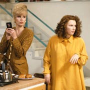 A tall white woman with blonde hair in bouffant updo standing beside a white man with curly dark hair in trendy kitchen