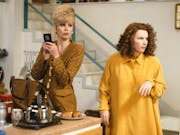 A tall white woman with blonde hair in bouffant updo standing beside a white man with curly dark hair in trendy kitchen