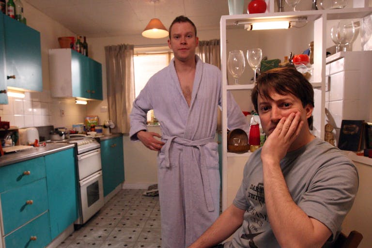 Two white men, one standing in a woman's dressing gown, the other sitting with his face in his hand