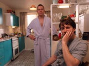 Two white men, one standing in a woman's dressing gown, the other sitting with his face in his hand
