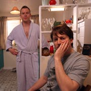 Two white men, one standing in a woman's dressing gown, the other sitting with his face in his hand