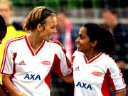 A young white woman and young British Asian woman in white and red football kits on a pitch looking jubilant