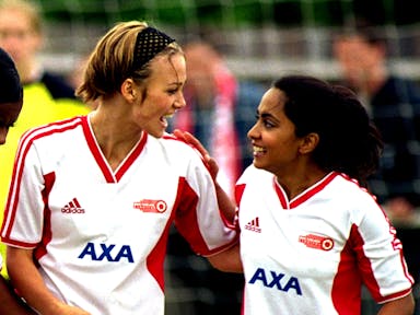 A young white woman and young British Asian woman in white and red football kits on a pitch looking jubilant 