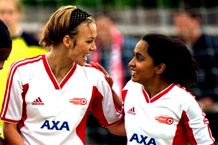 A young white woman and young British Asian woman in white and red football kits on a pitch looking jubilant