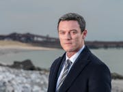 White man with short hair looks intently through the camera. In the background a beach with a destroyed pier