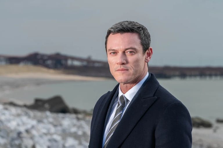 White man with short hair looks intently through the camera. In the background a beach with a destroyed pier