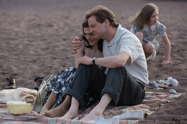 A white man and a white woman are sitting in an embrace on a blanket on the beach. A young girl is playing in the background.