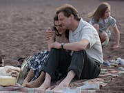 A white man and a white woman are sitting in an embrace on a blanket on the beach. A young girl is playing in the background.