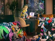 A white blonde woman sitting at a desk looking at a shopping page on a laptop. She is surrounded by piles of clothes and shoes.