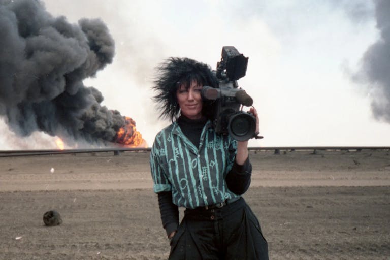 White woman holding a large broadcast camera looks are us smiling. Behind her a fire with heavy black smoke