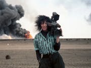 White woman holding a large broadcast camera looks are us smiling. Behind her a fire with heavy black smoke