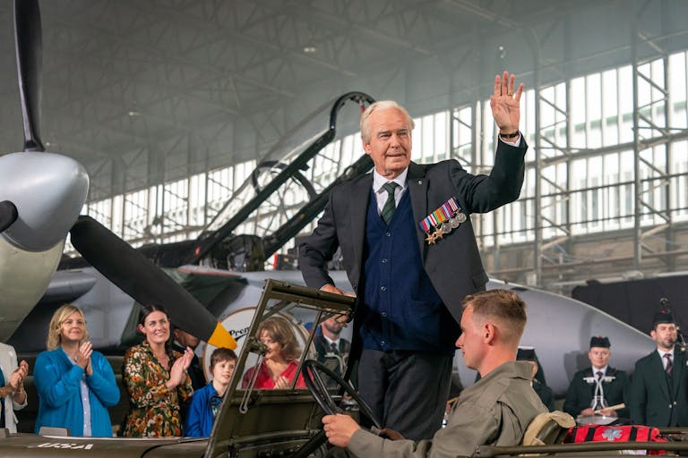 White elderly man, standing in a military car, in front of an old war plane, waives at an audience