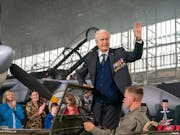 White elderly man, standing in a military car, in front of an old war plane, waives at an audience