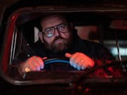 White man with thick rimmed-glasses, sitting behind the wheel in a black car