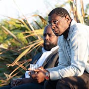 Two black men sitting near greenery are looking carefully towards the left edge of frame