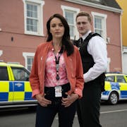Two police officers, a man and a woman looking at the camera stand in front of  two police cars