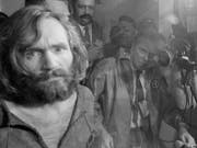 Black and white photo of a white man with long hair and a beard looking through the camera s reports surround him