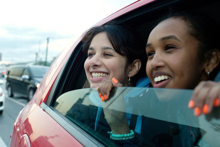 Two young girls smile as they look out of an open window from a moving car