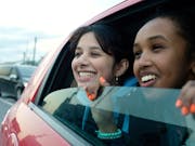 Two young girls smile as they look out of an open window from a moving car