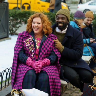 A white woman with short orange hair wearing a fuchsia pink coat sits beside a Black man in dark winter clothes and khaki woollen hat