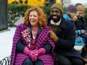 A white woman with short orange hair wearing a fuchsia pink coat sits beside a Black man in dark winter clothes and khaki woollen hat