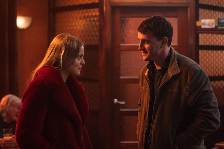 A young white woman with ginger hair stands opposite a young white man, an orange glow over them