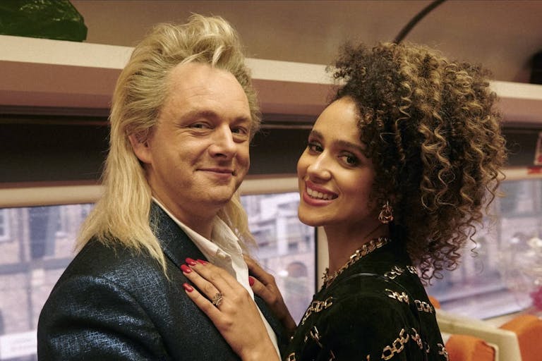 A middle aged white man with long blonde mullet in a train carriage with a mixed heritage young Black woman with her arms around him