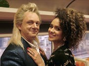 A middle aged white man with long blonde mullet in a train carriage with a mixed heritage young Black woman with her arms around him