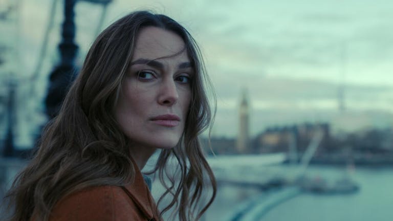 A white woman with long brown hair wearing a brown jacket looking serious and windswept standing beside the River Thames with Big Ben in the background 