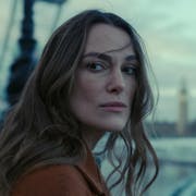 A white woman with long brown hair wearing a brown jacket looking serious and windswept standing beside the River Thames with Big Ben in the background